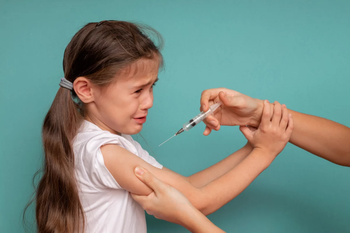 A distressed young girl resists as an adult holds her arm and approaches with a syringe.