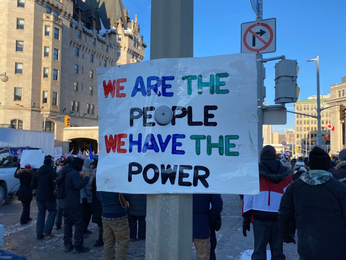 A protest sign reading “We are the people, we have the power” stands before a crowd gathered on a snowy Canadian street.