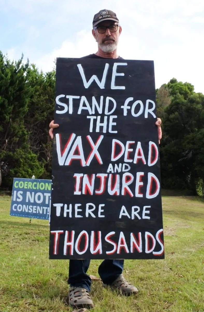 A bearded man standing outdoors holds a large sign reading, “We stand for the vax dead and injured—there are thousands.” A smaller sign behind him reads, “Coercion is not consent!”