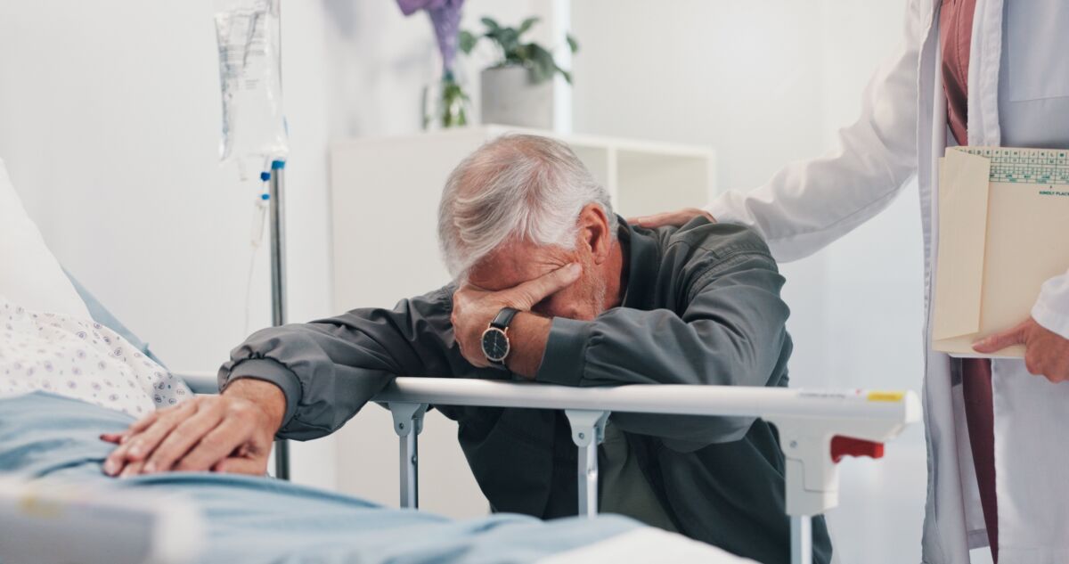 A distressed older man holds a patient’s hand beside a hospital bed as a doctor rests a comforting hand on his shoulder.