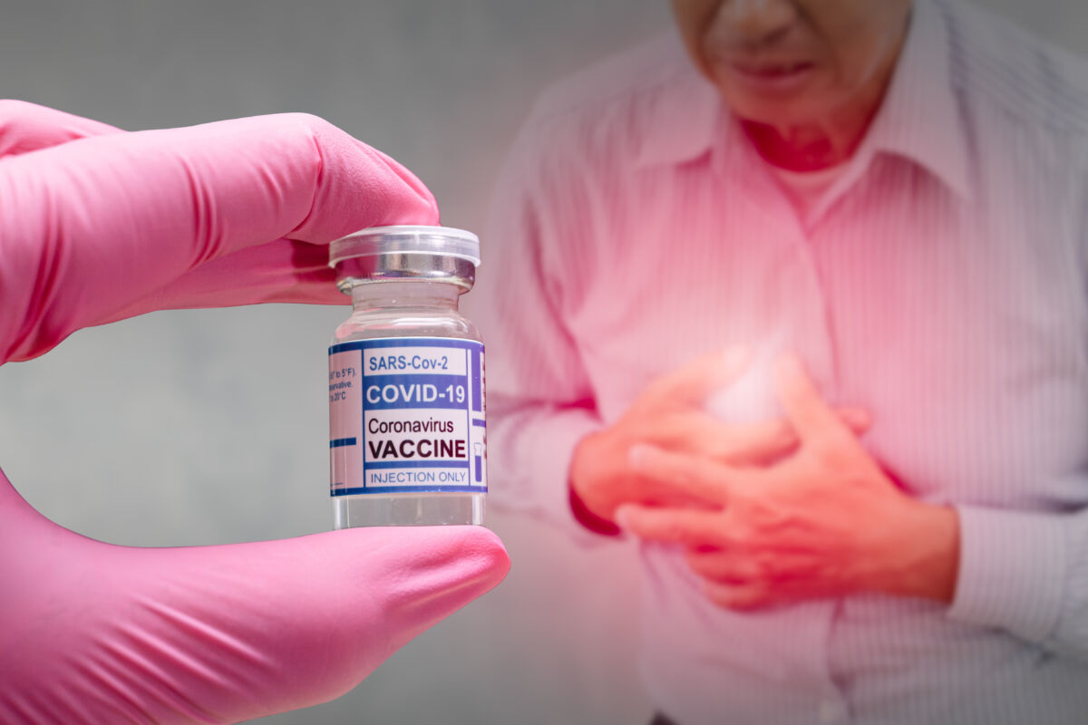 Gloved hand holding a vial labelled “COVID-19 vaccine” in the foreground, while a blurred man clutches his chest in the background.